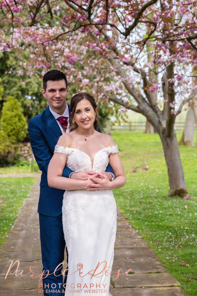 Photo of groom embracing his bride under cheery blossom treeon their wedding day in Milton Keynes