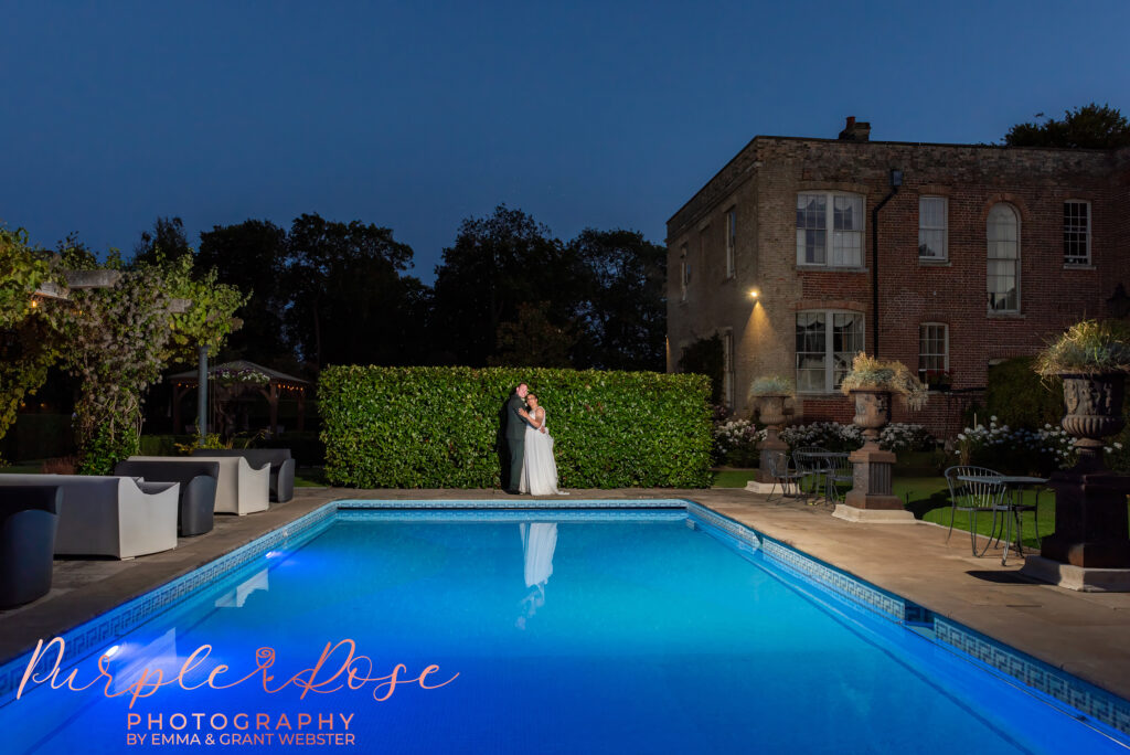 Photo of a bride and groom at night, stood by a swimming pool on their wedding day.