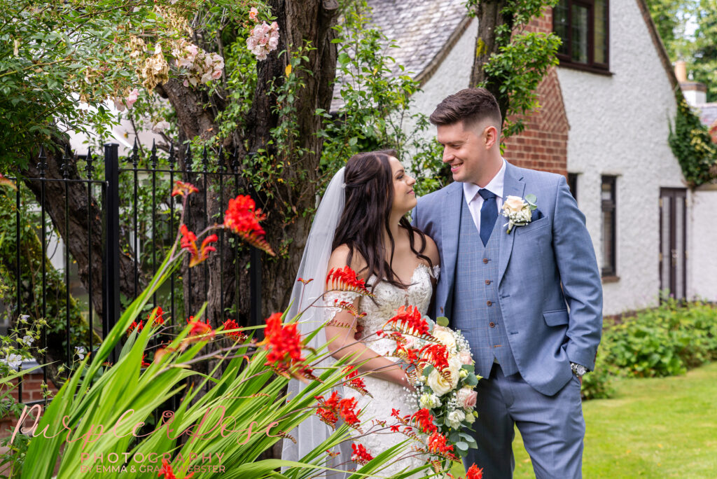 Bride and groom stood behind red flowers in a garden on their wedding day in Northampton