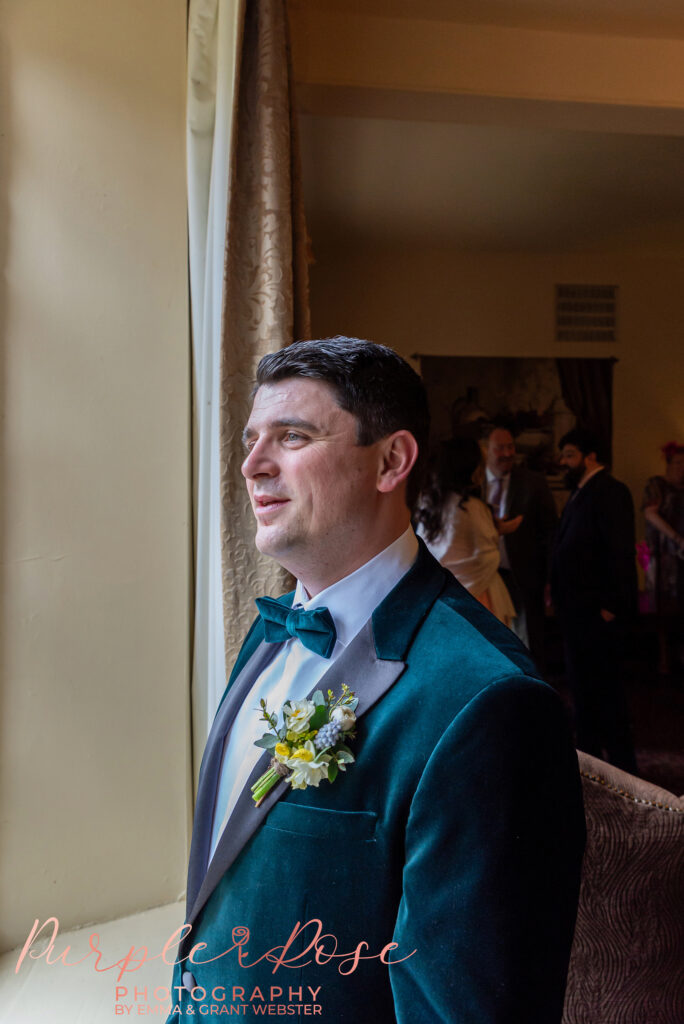 Photo of a groom in a green tuxedo looking out a window on his wedding morning in Northampton