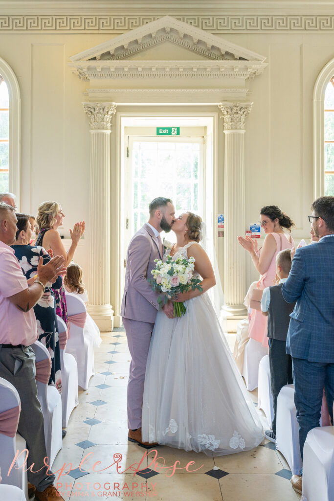 Bride and groom pausing to kiss as they leave their wedding ceremony at Chicheley Hall in Milton Keynes