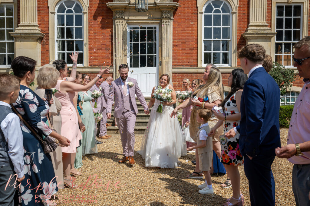 Bride and groom laughing as they leave their wedding ceremony while their guest throw confetti at Chicheley Hall in Milton Keynes