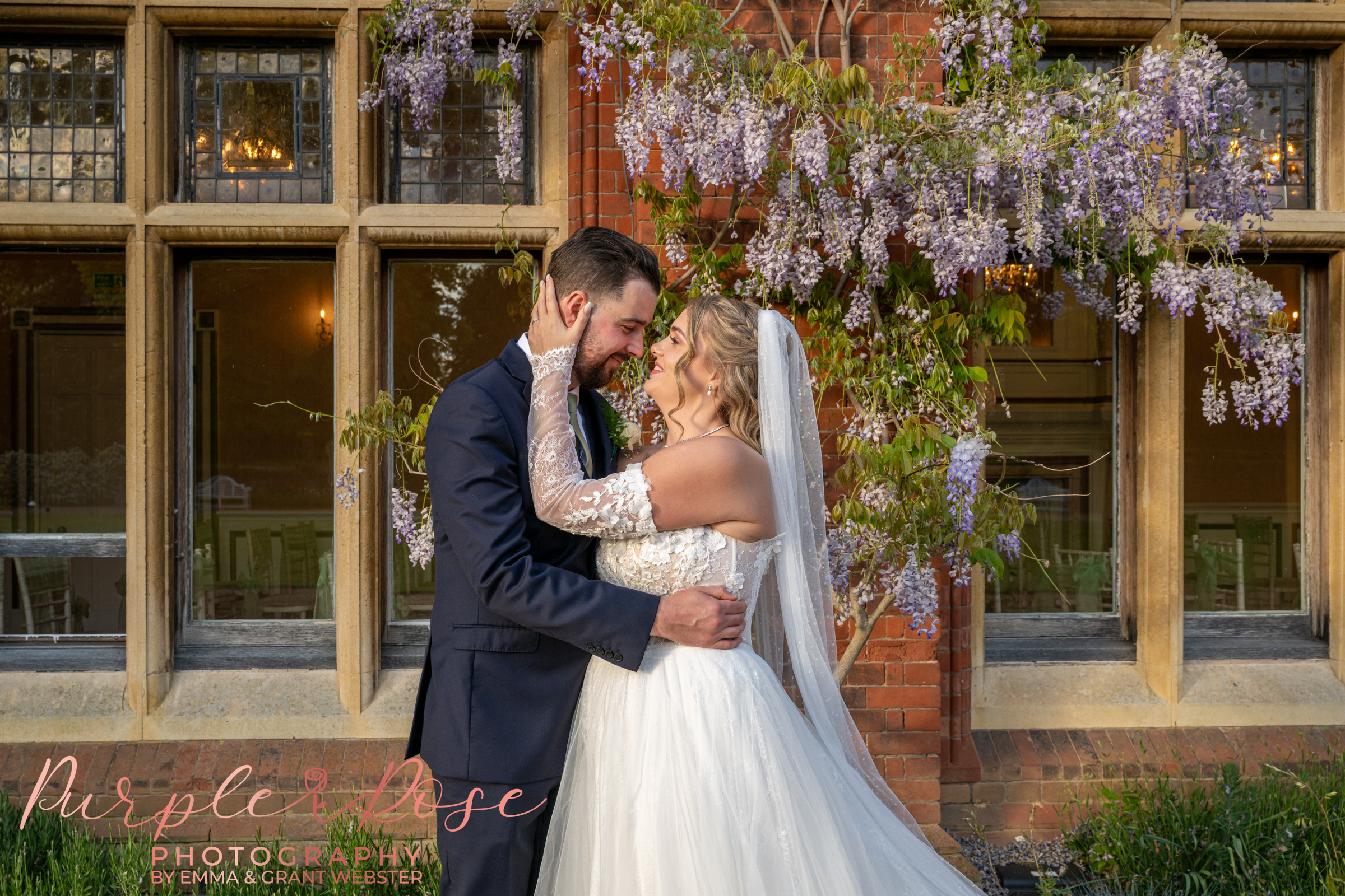 Photo of a bride and groom stood outside their wedding venue surrounded by lilac wisteria blooms