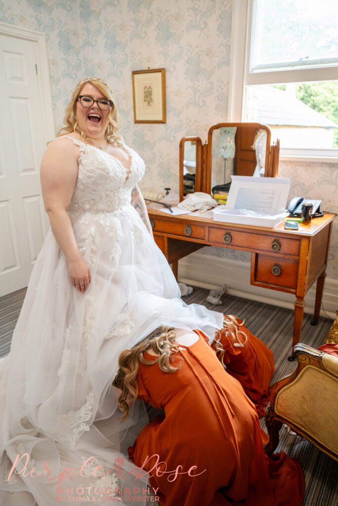 Bride laughing while her bridesmaid are helping her get her wedding shoes on